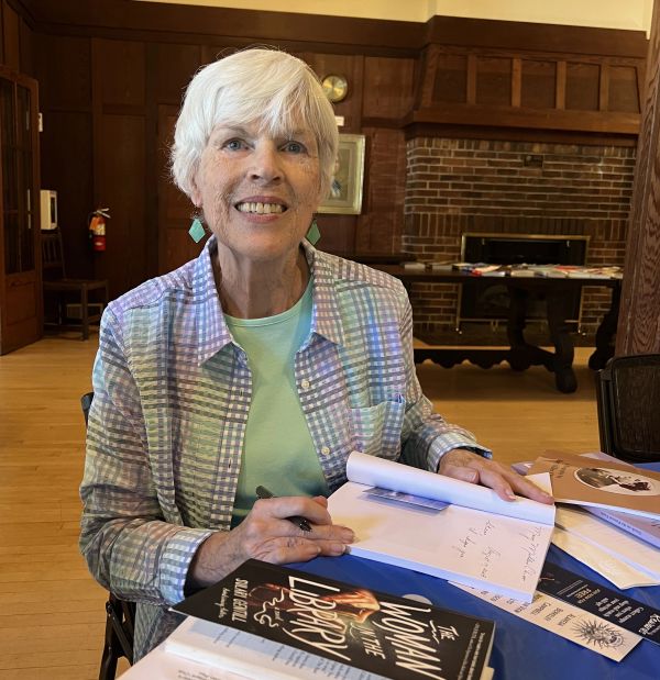 Elderly woman signing books at a table in a cozy room.