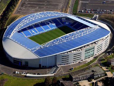 Aerial view of a large modern football stadium with a blue roof and green pitch.