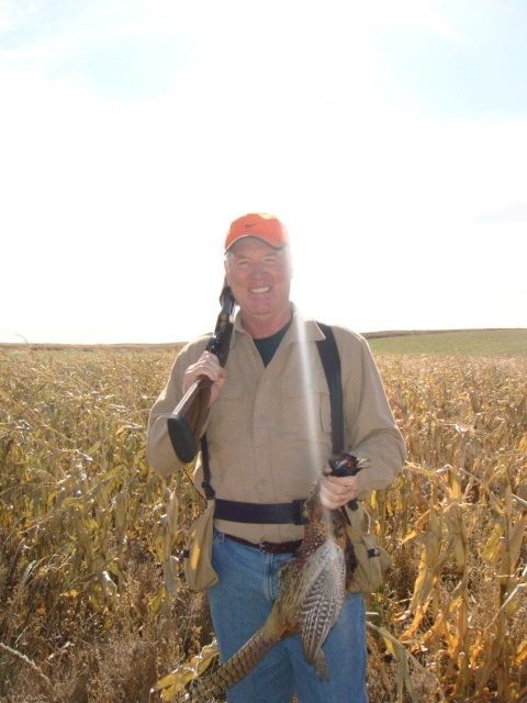 Man holding a hunting rifle and a pheasant in a field.