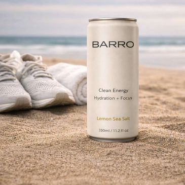 A can of BARRO Lemon Sea Salt drink on a sandy beach with shoes and towel in the background.