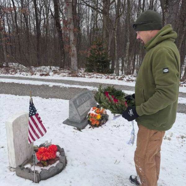 A man in winter clothing holding a wreath at a snowy gravesite with an American flag.
