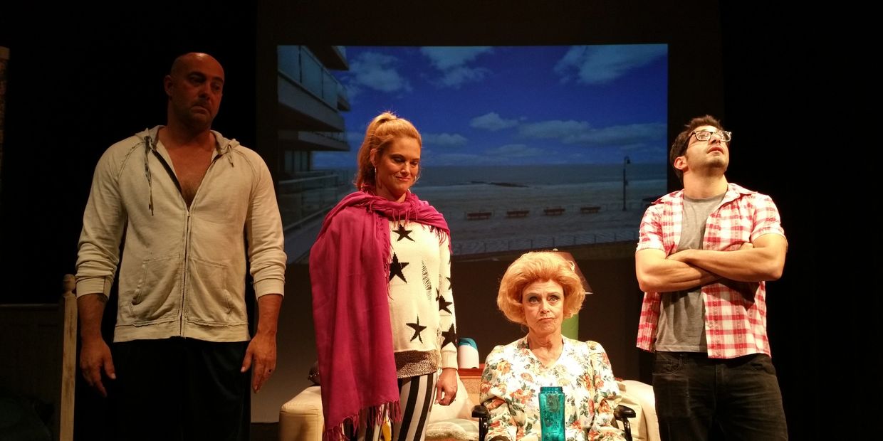 Four actors on stage with a beach backdrop, portraying a dramatic scene.