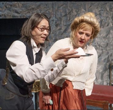 Two actors in vintage costumes examine a plate with a white substance on stage.