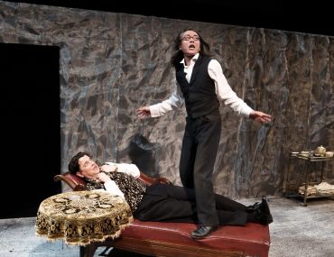 Two actors perform on stage, one lying on a chaise lounge, the other standing.