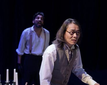 Two men in vintage clothes acting intensely on stage with candles in foreground.