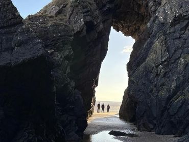 Three people walking on a beach through a large natural rock arch.