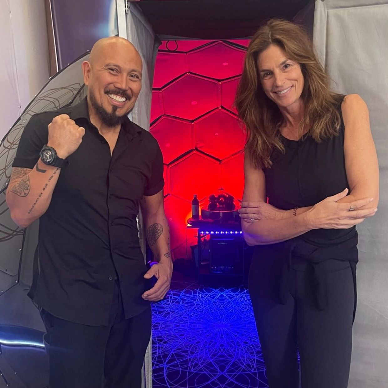 Two smiling people posing in front of a brightly lit, geometric-patterned backdrop.