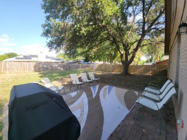 Backyard patio with chairs and a grill under a large tree.