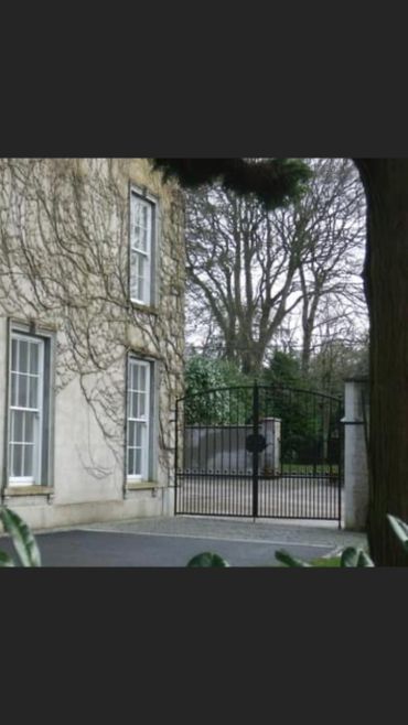 Stone building with leafless vines next to a closed metal gate and trees.