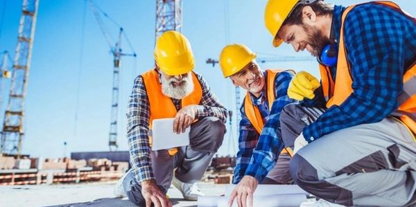 Three construction workers in hard hats and vests reviewing blueprints on site.