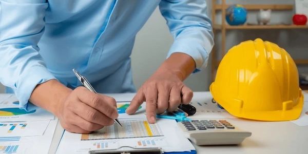 Person reviewing construction documents with calculator and yellow hard hat nearby.
