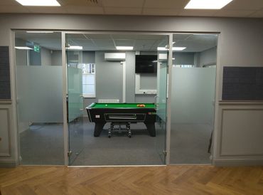 A pool table behind glass doors in a modern room with gray walls and wood flooring.
