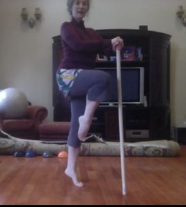 Woman practicing balance exercise with a wooden stick in a living room.