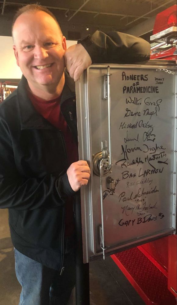 A man smiles beside a signed metal panel titled 'Pioneers of Paramedicine.'