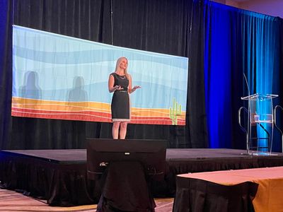 Julia Myers speaking from a large ballroom stage wearing a black Clavin Klein dress