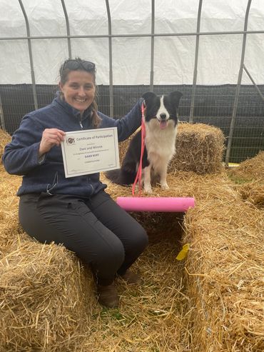 Woman and dog with barn hunt certificate among hay bales.