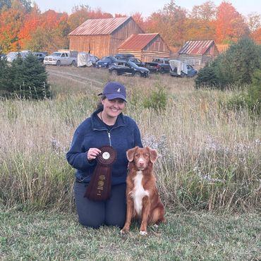 Woman kneeling with a dog holding a prize ribbon outdoors.