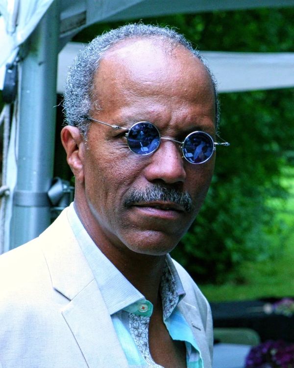 Man wearing round blue-tinted sunglasses and a light-colored suit jacket.