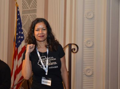 Woman wearing a volunteer badge poses with a fist near an American flag indoors.