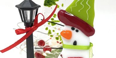 A fused glass snowman standing next to a lamppost in front of a decorated glass tree.