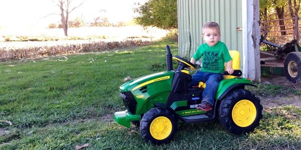 A young boy sits on a green and yellow toy tractor outside.