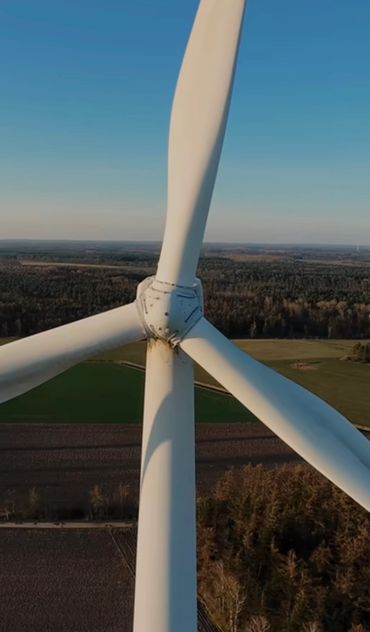 Close-up of a wind turbine with rust near the hub against a forest and field background.