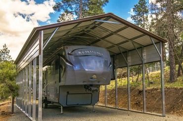 A large metal carport sheltering a Komfort RV in a wooded area.