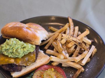 Cheeseburger with guacamole, lettuce, tomato, pickles, and seasoned fries on a black plate.