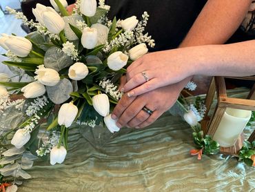 Bride and groom hold hands over their bouquet showing their wedding rings.