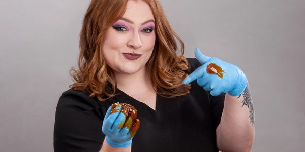 A redhead woman with tattoos wearing vibrant makeup points to the sugaring paste on the other hand