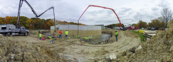 Construction workers pour concrete into a large circular foundation using concrete pump trucks.