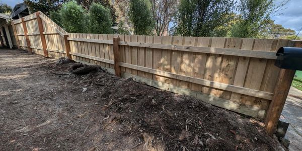 A wooden fence runs along a dirt pathway in a backyard.