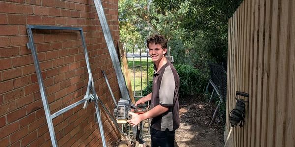 Young man using a power tool outdoors near a brick wall and wooden fence.