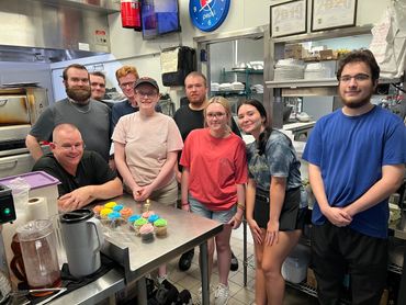 A group of people posing in a kitchen with colorful cupcakes on the table.