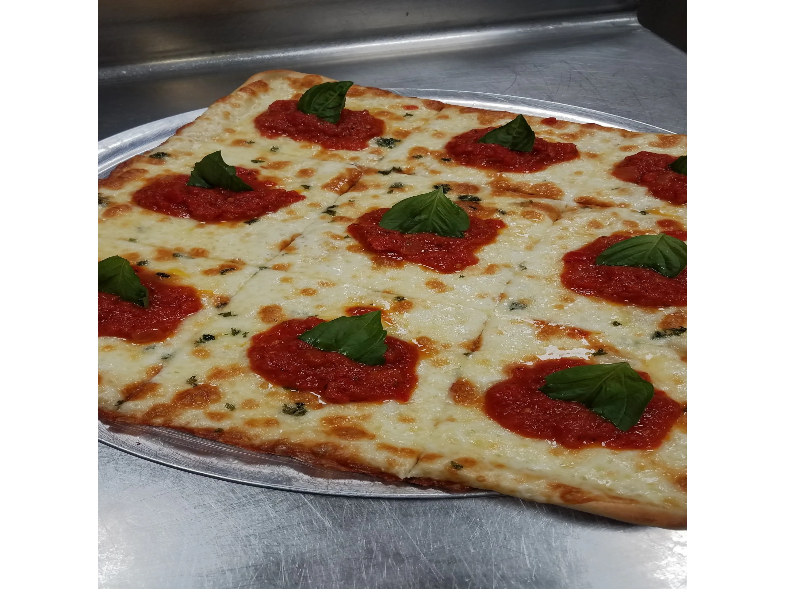 Rectangular pizza with cheese, tomato sauce dollops, and basil leaves on a metal tray.