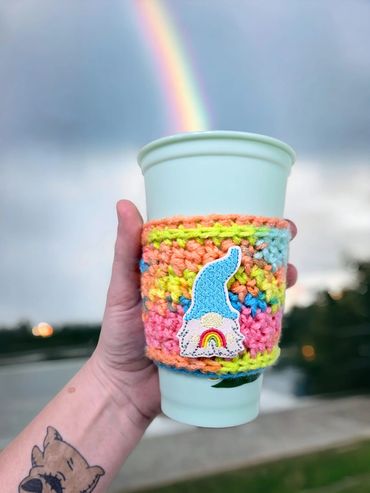 Hand holding a cup with a colorful knitted sleeve and a rainbow patch, rainbow in the background.