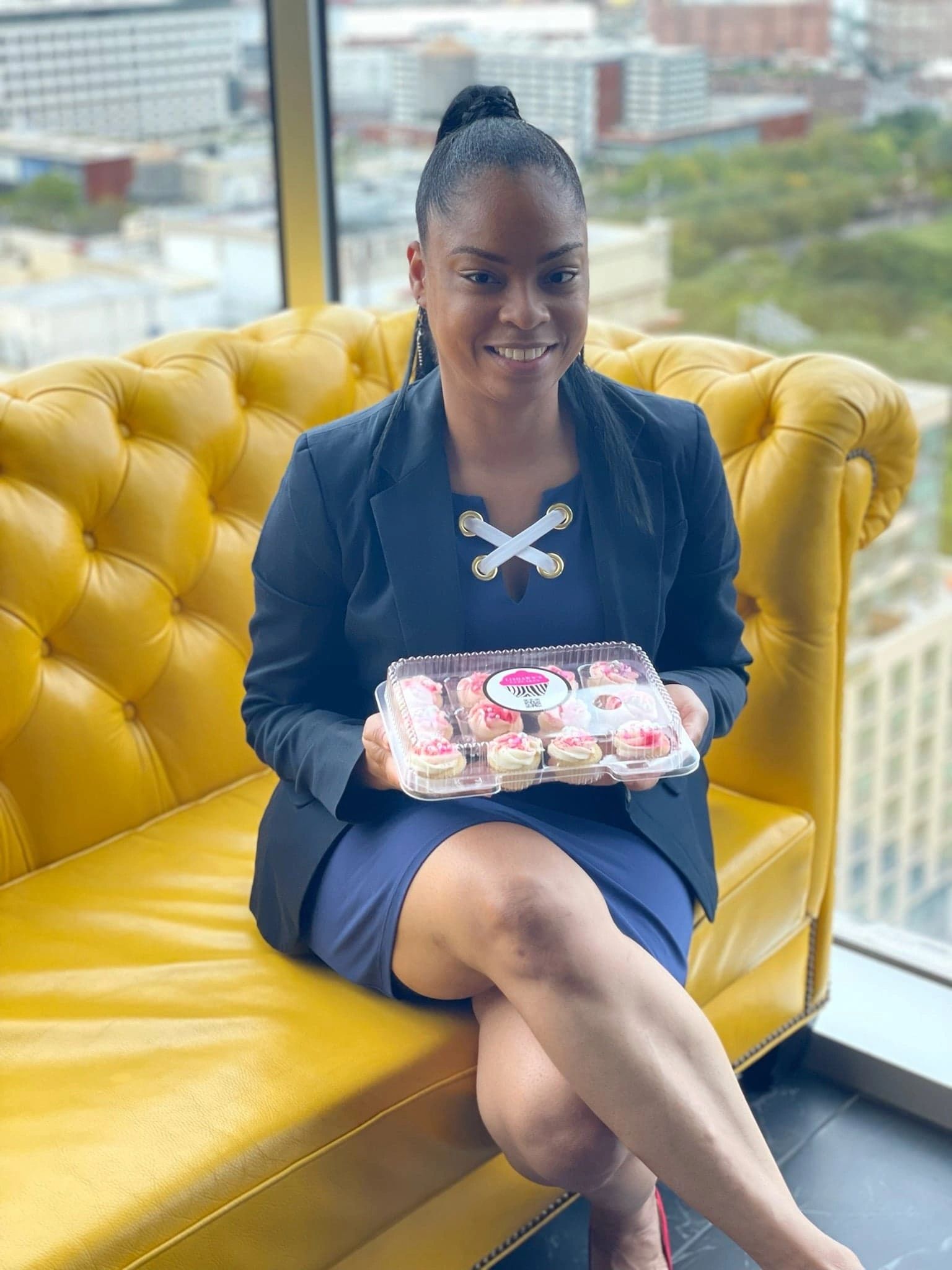 Lishawn holding a dozen of mini cupcakes on a yellow sofa with the Brooklyn skyline in the back