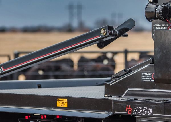Close-up of a black HydraBed 3250 trailer with caution sign and cattle in the background.