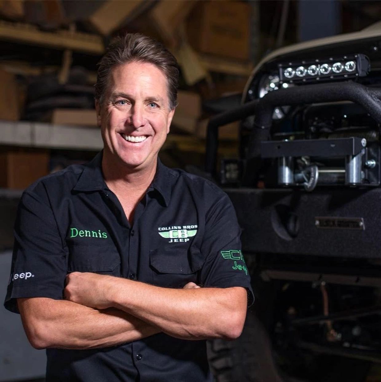 A smiling man named Dennis in a Collins Bros Jeep shirt stands with arms crossed by a Jeep.