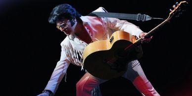 Elvis Presley in a dynamic stage pose with guitar, wearing a white jumpsuit.