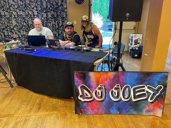 Three people at a DJ booth with speakers and a sign saying "DJ JOEY"