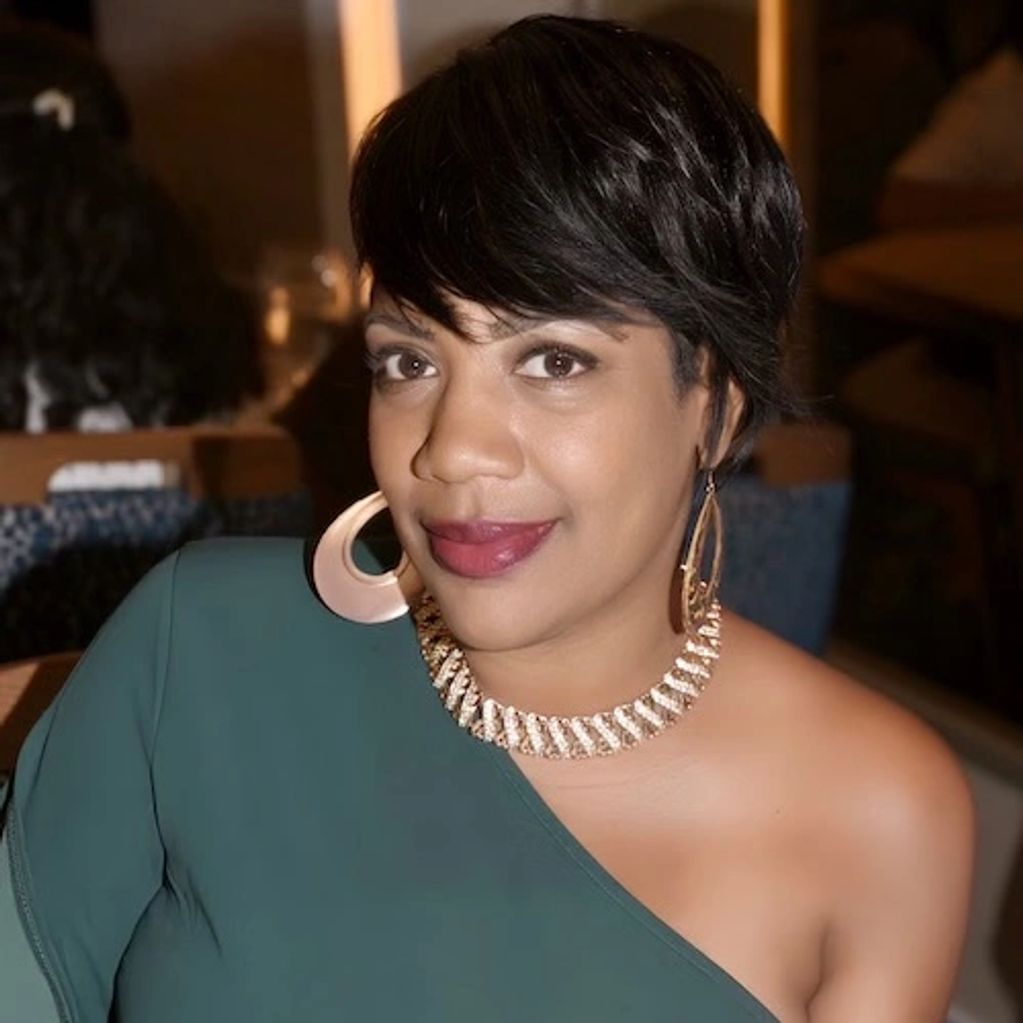 Woman with short black hair wearing a green one-shoulder dress and statement jewelry.