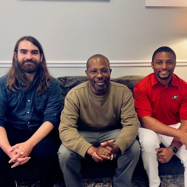 Three men sitting side by side on a couch, smiling at the camera.