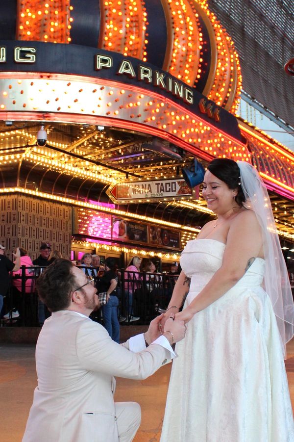 A man proposes to a woman in a wedding dress under bright neon lights.