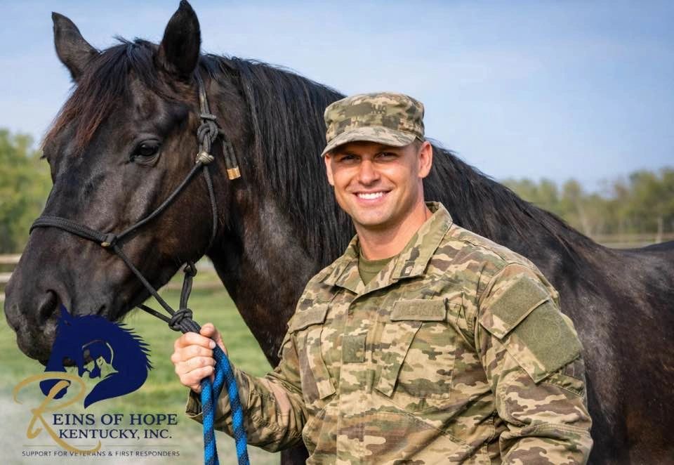 A smiling soldier in camouflage holding a black horse's reins outdoors.