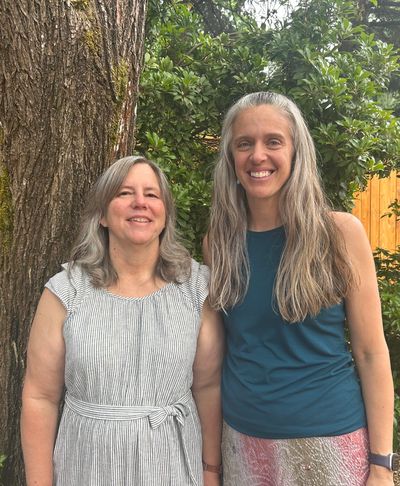 Two smiling women standing outdoors in front of a tree and greenery.
