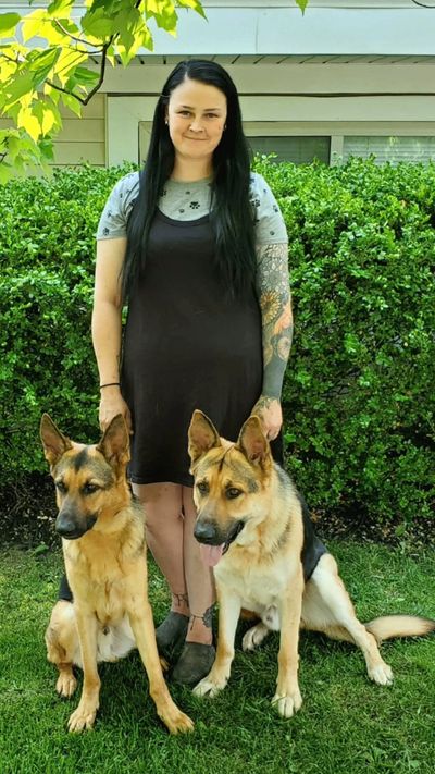 Tia Livengood stands outdoors on grass in front of green bushes, holding two German Shepherds seated
