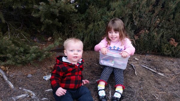 Two young children sitting outdoors among trees and dirt.