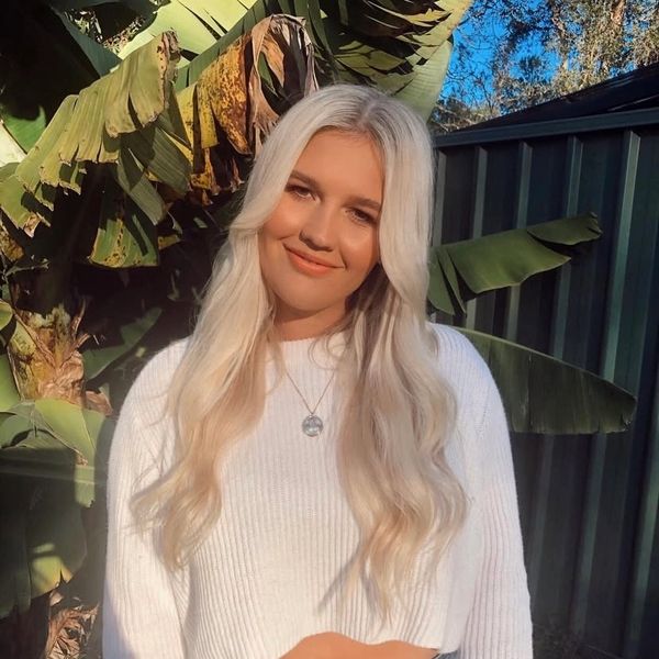 Young woman with long blonde hair in a white sweater and jeans, smiling outdoors.
