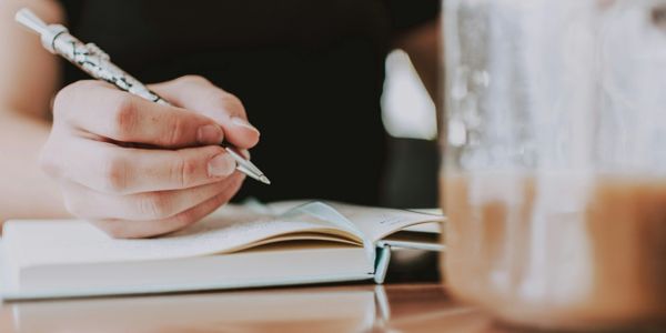 Person writing in a notebook with a pen beside a glass of coffee.